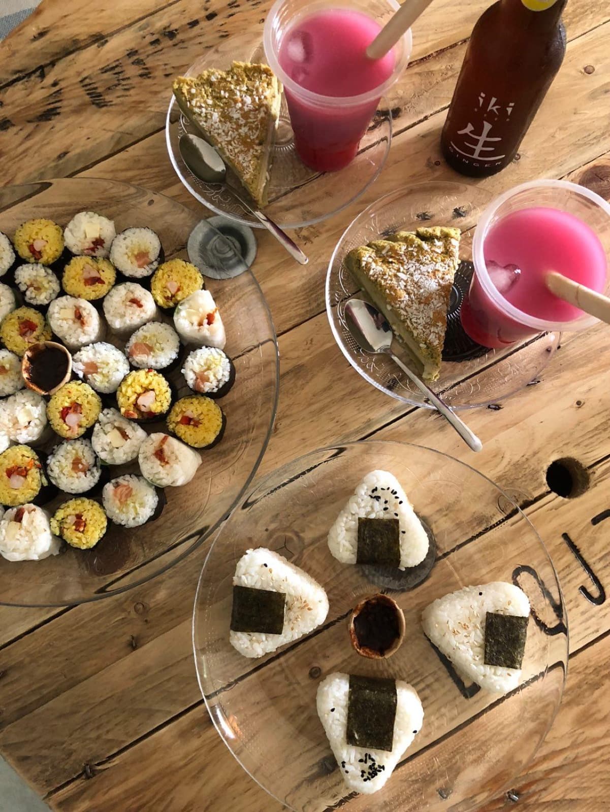 Table en bois garnie de plateaux de makis et d’onigiris, de parts de gâteau et de deux bubble tea roses.