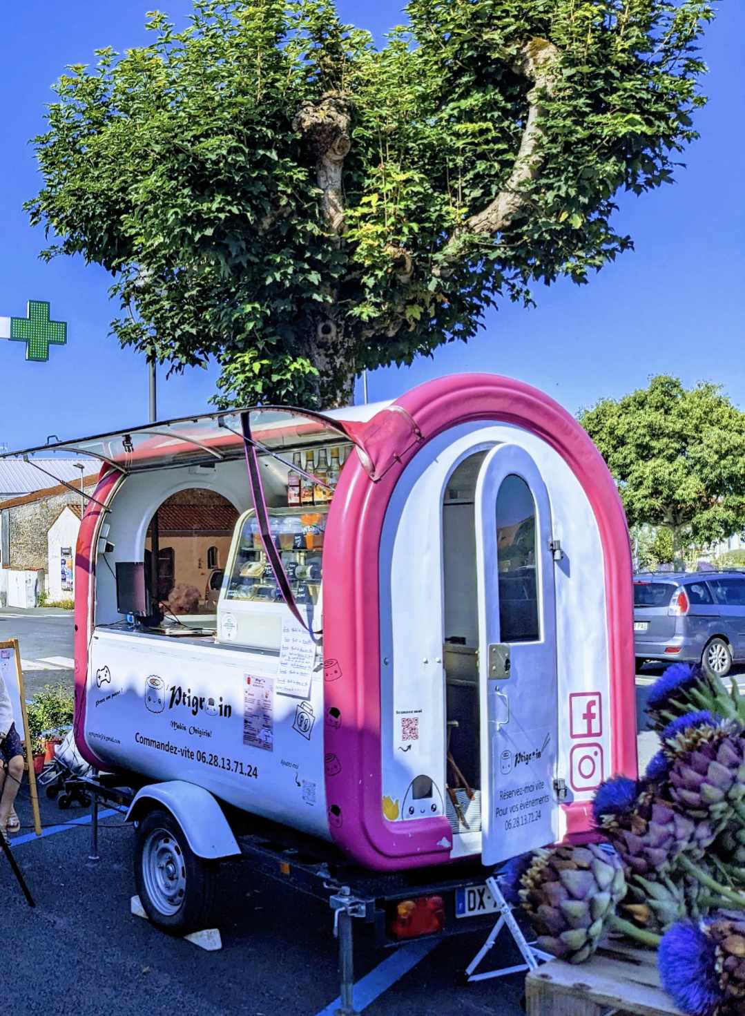 La roulotte O’ptigrain installée sur un marché sous un arbre, par grand soleil.